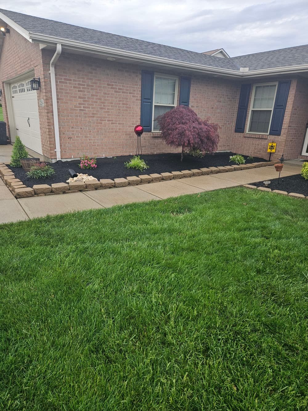 Front yard with green grass, brick house, flower bed, and ornamental red garden decoration.