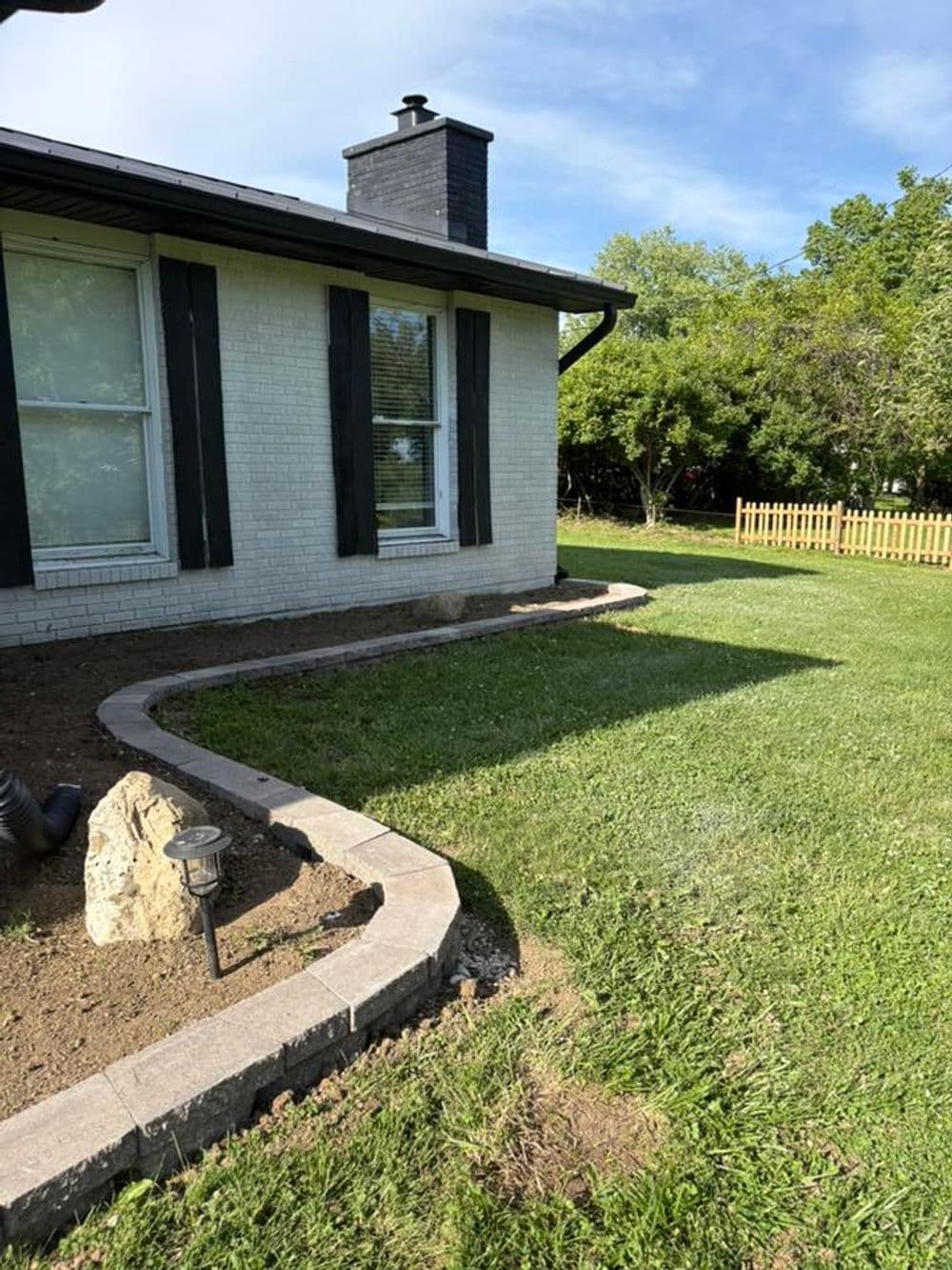 Side view of a house with landscaping, a chimney, and a fenced yard in sunlight.