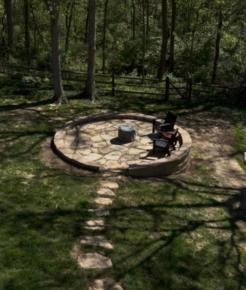 Stone fire pit surrounded by grassy area and chairs in a wooded backyard setting.