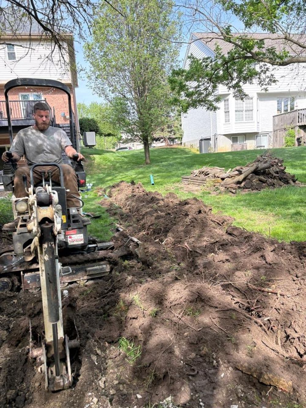 Man using a trenching machine in a residential yard, with trees and a house in the background.