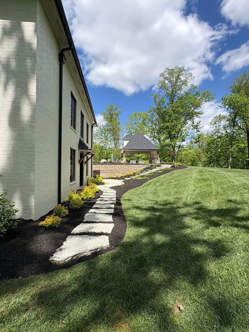 Modern home exterior with landscaped path, lush greenery, and a gazebo in a sunny setting.